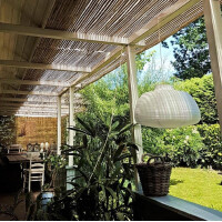 Rollzaun aus Bambus bietet natürlichen Sichtschutz auf einer Terrasse mit weißen Holzpfosten und Dach. Der Zaun erzeugt eine gemütliche, privat anmutende Atmosphäre und fügt sich harmonisch in die grüne Umgebung ein.