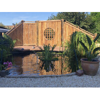 Sichtschutz aus Bambus in einem asiatisch inspirierten Garten, der ein ruhiges Ambiente schafft. Das Zaunelement ANJI NIGRA verbindet natürliche Ästhetik mit Funktionalität und bietet Privatsphäre durch seine dichten, vertikalen Bambusstäbe. Ein kreisförmiges Fenster mit Gittermuster im Zentrum des Zauns sorgt für einen dekorativen Akzent. Der Bambuszaun steht neben einem kleinen Teich, der die friedliche Atmosphäre unterstreicht.