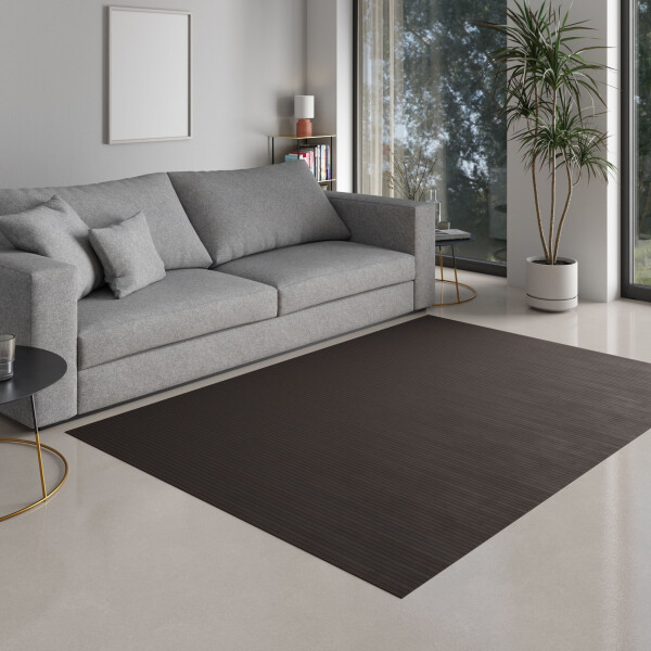 Kleines Mädchen sitzt auf einem dunklen Bambusteppich im Wohnzimmer und malt. Der Teppich hat eine elegante, dunkle Farbe und passt sich harmonisch in die moderne Einrichtung ein. Der Raum ist hell und freundlich gestaltet, mit einem grauen Sofa und grünen Pflanzen im Hintergrund.