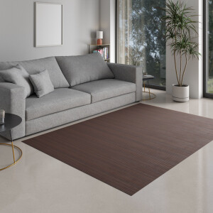 Mädchen sitzt auf einem mochafarbenen Bambusteppich im Wohnzimmer, zeichnet auf Papier und lächelt in die Kamera. Der Teppich hat eine moderne, gestreifte Textur und passt perfekt in das helle, einladende Ambiente mit Sofa und Pflanzen.