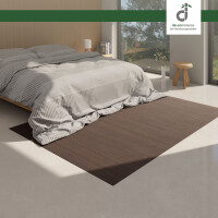 Modernes Wohnzimmer mit einem dunkelbraunen Bambusteppich auf hellem Laminatboden. Ein moderner Sessel und ein Sideboard mit Dekoration ergänzen die Szene. Große Fenster lassen natürliches Licht herein, und ein stilvoller Wandleuchter hängt über dem Teppich.