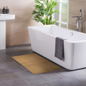 Modernes Badezimmer mit freistehender, rechteckiger Badewanne neben einem Fenster und einer grünen Zimmerpflanze. Ein graues Handtuch hängt über dem Wannenrand. Auf dem dunklen Fliesenboden liegt eine natürliche Bambus-Badematte, links ist ein Waschbecken mit Pflegeprodukten zu sehen.