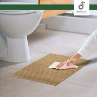 Eine Hand reinigt eine natürliche Bambus-Badematte mit einem Tuch vor einer weißen Toilette auf hellem Fliesenboden in einem modernen badezimmer, oben rechts das DE-COmmerce Logo.