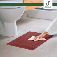 Eine Hand reinigt eine warm-rote Bambus-Badematte mit einem Tuch vor einer weißen Toilette auf hellem Fliesenboden in einem modernen badezimmer, oben rechts das DE-COmmerce Logo.