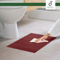 Badteppich LINES Wunschmaß aurora in dunkelrotem Bambusdesign auf weißem Badezimmerboden. Eine Person reinigt den Teppich mit einem weißen Tuch, während im Hintergrund eine weiße Toilette sichtbar ist.