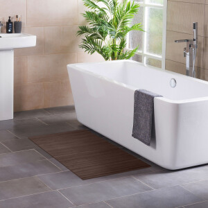 Modernes Badezimmer mit freistehender, rechteckiger Badewanne neben einem Fenster und einer grünen Zimmerpflanze. Ein graues Handtuch hängt über dem Wannenrand. Auf dem dunklen Fliesenboden liegt eine Kaffeebraune Bambus-Badematte, links ist ein Waschbecken mit Pflegeprodukten zu sehen.