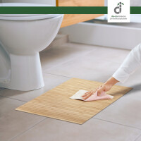 Eine Hand reinigt eine helle Bambus-Badematte mit einem Tuch vor einer weißen Toilette auf hellem Fliesenboden in einem modernen badezimmer, oben rechts das DE-COmmerce Logo.