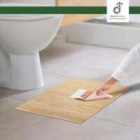 Eine Person reinigt einen Bambus-Badteppich mit einem Tuch neben einer Toilette im Badezimmer. Der Teppich besteht aus natürlichen Bambuslamellen und bietet eine moderne, minimalistische Optik.