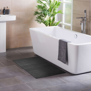 Modernes Badezimmer mit freistehender, rechteckiger Badewanne neben einem Fenster und einer grünen Zimmerpflanze. Ein graues Handtuch hängt über dem Wannenrand. Auf dem dunklen Fliesenboden liegt eine stahlgraue Bambus-Badematte, links ist ein Waschbecken mit Pflegeprodukten zu sehen.