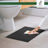 Eine Hand reinigt eine stahlgraue Bambus-Badematte mit einem Tuch vor einer weißen Toilette auf hellem Fliesenboden in einem modernen badezimmer, oben rechts das DE-COmmerce Logo.