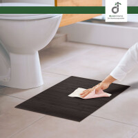 Eine Hand reinigt eine dunkle schwarzbraune Bambus-Badematte mit einem Tuch vor einer weißen Toilette auf hellem Fliesenboden in einem modernen badezimmer, oben rechts das DE-COmmerce Logo.