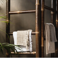 Dunkler Bambus-Raumteiler mit mehreren Querstreben, der als dekoratives Element dient. Ein wolliges Kissen und ein Handtuch sind daran drapiert, was eine gemütliche Atmosphäre schafft. Grüne Pflanzen im Hintergrund ergänzen den natürlichen Look.