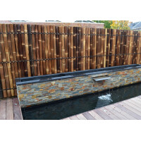 Bambus Sichtschutz XXL SLIM als dekoratives Zaunelement in Naturfarbe, dicht aneinandergereihte Bambusstäbe, oberer Rand mit stabiler Abgrenzung, hinter einem modernen Wasserfall aus Stein.