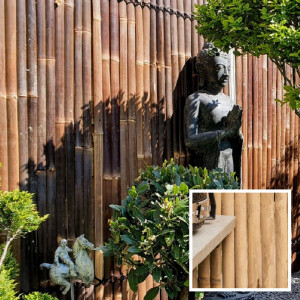 Dunkler Kisu Bambuszaun („Nigra“) im Garten, hinter einer Skulpturfigur, umgeben von üppigen Pflanzen, vermittelt eine elegante und natürliche Atmosphäre.