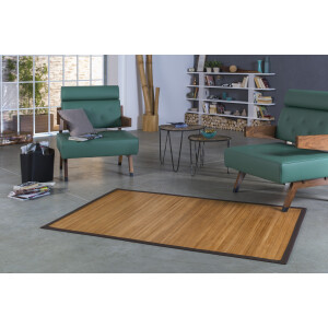 Großer, rechteckiger Bambusteppich mit charakteristischen 17mm breiten Stegen in natürlicher Holzoptik. Der Teppich ist von einer eleganten, braunen Bordüre umgeben und wird in einem modernen Wohnzimmer präsentiert, das durch grüne Sessel und minimalistische Dekoration besticht. Die natürliche Ästhetik des Bambus harmoniert mit dem urbanen Stil des Raumes.