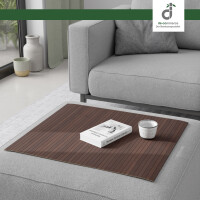 Flexible Couchablage aus Bambus mit Stegstruktur in mittelbraun mit Kaffeeton liegt auf einer grauen Couch und dient als stabile Ablagefläche für Buch und Kerze – ideal als Sofa-Tablett oder Schutz für Polster, oben rechts DE-COmmerce Logo.