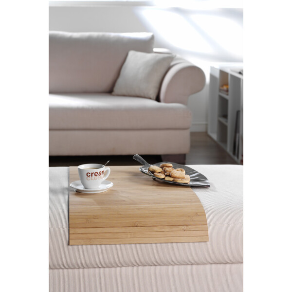 Elegante Couchablage aus Bambus in Naturfarbe, individuell anpassbar, mit einer Tasse Kaffee und einem Teller Gebäck darauf, platziert vor einem hellen Sofa in einem modernen Wohnzimmer.