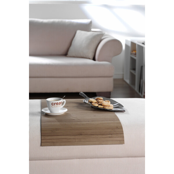 Elegante Couchablage aus Bambus im Taupe-Design, die stilvoll auf einem Sofa platziert ist. Darauf stehen ein Kaffeebecher mit der Aufschrift cream und ein Teller mit Gebäck. Die Ablage verbindet natürliche Ästhetik mit Funktionalität.