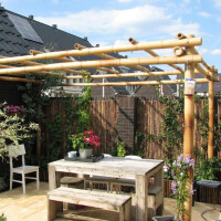 Bambus Pergola mit robusten Bambusstangen, die ein stabiles Dach bilden. Ideal für Garten oder Terrasse, bietet Schatten und eine natürliche Atmosphäre. Kombiniert mit rustikaler Holzmöbelgarnitur und Pflanzen.