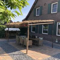 Bambus Pergola mit natürlichem Strohdach, die Schatten spendet über einer Sitzecke mit Gartenmöbeln vor einem Haus mit dunkelgrüner Fassade.