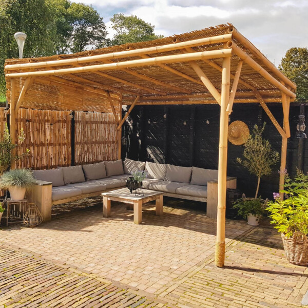 Eine große Bambus-Pergola mit natürlichem Dach aus Guadua-Bambus, die eine gemütliche Sitzecke mit Sofa und Couchtisch im Garten beschattet. Die Pergola bietet einen stilvollen Rückzugsort im Freien, ideal für entspannte Stunden in einer grünen Umgebung. Die Maße der Pergola betragen 200x300x300 cm.