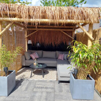 Bambus Pergola Guadua mit natürlichem Strohdach und Bambuswänden, die Schatten und Privatsphäre bieten. Eine gemütliche Sitzecke mit Sofa und Couchtisch darunter, umgeben von Pflanzen in Betonkübeln.