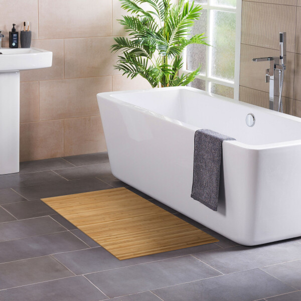 Modernes Badezimmer mit freistehender, rechteckiger Badewanne neben einem Fenster und einer grünen Zimmerpflanze. Ein graues Handtuch hängt über dem Wannenrand. Auf dem dunklen Fliesenboden liegt eine warm gelbe Bambus-Badematte, links ist ein Waschbecken mit Pflegeprodukten zu sehen.