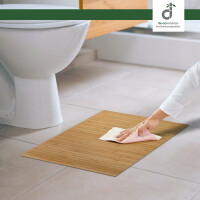Eine Hand reinigt eine warm gelbe Bambus-Badematte mit einem Tuch vor einer weißen Toilette auf hellem Fliesenboden in einem modernen badezimmer, oben rechts das DE-COmmerce Logo.