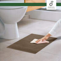 Eine Hand reinigt eine hellgrau-braune Bambus-Badematte mit einem Tuch vor einer weißen Toilette auf hellem Fliesenboden in einem modernen badezimmer, oben rechts das DE-COmmerce Logo.