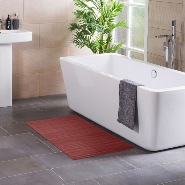 Modernes Badezimmer mit freistehender, rechteckiger Badewanne neben einem Fenster und einer grünen Zimmerpflanze. Ein graues Handtuch hängt über dem Wannenrand. Auf dem dunklen Fliesenboden liegt eine warm-rote Bambus-Badematte, links ist ein Waschbecken mit Pflegeprodukten zu sehen.
