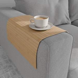 Helle Bambus-Couchablage mit flexiblen Lamellen liegt über der Armlehne eines grauen Sofas und dient als stabile Ablagefläche für eine Tasse Kaffee auf Untertasse.
