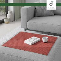 Flexible Couchablage aus Bambus mit Stegstruktur in einem warmen Rot liegt auf einer grauen Couch und dient als stabile Ablagefläche für Buch und Kerze – ideal als Sofa-Tablett oder Schutz für Polster, oben rechts DE-COmmerce Logo.