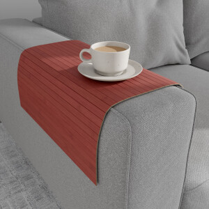 Bambus-Couchablage im warmen Rotton mit flexiblen Lamellen liegt über der Armlehne eines grauen Sofas und dient als stabile Ablagefläche für eine Tasse Kaffee auf Untertasse.