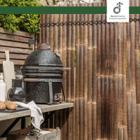 Outdoor-Küchenbereich mit dunklem Kisu Bambuszaun („Nigra“), Grillgerät auf Holzregal, Backwaren und Küchenutensilien, gemütliche Grillatmosphäre.