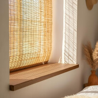 Sonnendurchflutetes Fenster mit heller, heruntergelassener Schilfjalousie. Auf der hölzernen Fensterbank fällt warmes, gefiltertes Licht ein, daneben steht eine Vase mit getrocknetem Pampasgras in einem gemütlichen Raum.