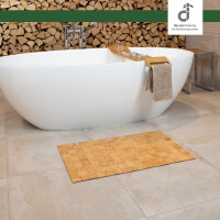 In einem modernen Badezimmer mit hellen Fliesen steht eine freistehende Badewanne mit Badewannentisch und Handtuch. Im Hintergrund ist Kaminholz und im Vordergrund liegt eine Bambus-Badematte in der Farbe Nature. Oben rechts ist das DE-COmmerce Logo.