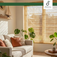 Helles, gemütlich eingerichtetes Wohnzimmer mit einem beigen Sofa. Vor dem großen Fenster hängen Schilfjalousien, durch die warmes Sonnenlicht fällt. Mehrere Monstera-Pflanzen stehen vor dem Fenster, und an der Wand hängt ein Regal mit Deko-Objekten. Oben rechts befindet sich das DE-COmmerce Logo.