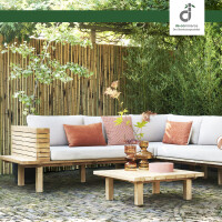 Gemütliche Gartensitzecke mit Holz-Loungesofa und hellen Polstern vor natürlichem Bambus-Windschutzzaun, umgeben von üppigen grünen Pflanzen; niedriger Holztisch mit Dekoration im Vordergrund, oben rechts das DE-COmmerce Logo.
