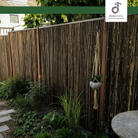 Moderner Garten mit dunklen Bambus-Windschutz an Metallpfosten, Holzsteg und Rasenfläche, im Hintergrund eine hohe grüne Hecke unter blauem Himmel, oben rechts das DE-COmmerce Logo.