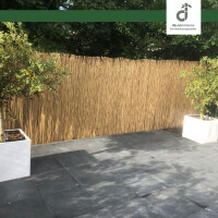 Terrasse mit dunklen Bodenfliesen und hohem Sichtschutz aus naturbelassener Schilfrohrmatte; links und rechts stehen weiße Pflanzkübel mit kleinen Bäumen, dahinter dichtes grünes Laub; oben rechts das DE-COmmerce Logo.