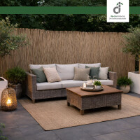 Outdoor-Lounge auf Terrasse mit grauen Steinplatten und hoher Schilfmatte als Sichtschutz; Rattan-Sofa mit hellen Polstern und grünen sowie beigen Kissen, Rattan-Couchtisch mit Tablett und große Laterne neben Topfpflanzen; oben rechts DE-COmmerce Logo.