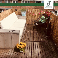 Gemütlicher Balkon mit Schilfmatte als Sichtschutz, Holzdielen und Lichterkette; Holzsofa mit weißen Polstern, schwarzer Korbsessel mit grünem Kissen und Glas mit Sonnenblumen im Vordergrund; oben rechts DE-COmmerce Logo.