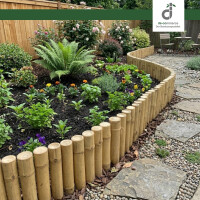 Gepflegtes Gartenbeet mit vielfältigen Blumen und Kräutern, eingefasst von einer geschwungenen Bambus-Beeteinfassung; daneben verläuft ein Steinweg mit Kies, im Hintergrund ein Holzzaun und Sitzbereich; oben rechts das DE-COmmerce Logo.