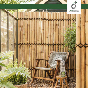 Sitzecke mit einem Sichtschutzelelement aus vertikal gebundenen Bambusspalten; davor steht ein Holzstuhl mit geflochtener Sitzfläche, umgeben von grünen Pflanzen; oben rechts das DE-COmmerce Logo.