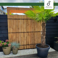 Garten- bzw. Terrassenbereich mit einer Sichtschutzwand aus hellen, vertikal mit Palmseil gebundenen Bambuslatten, kombiniert mit dunklen Holzelementen; davor stehen dekorative Pflanzkübel und eine große Palme in einem Topf. Oben rechts ist das DE-COmmerce Logo.
