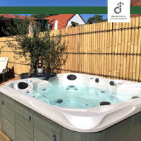 Whirlpool im Garten mit klarem Wasser, umgeben von einer natürlichen Bambus-Sichtschutzwand; daneben ein kleiner Tisch mit Weinflasche und zwei Gläsern sowie ein Olivenbaum im Topf, im Hintergrund ein Haus mit rotem Ziegeldach unter blauem Himmel; oben rechts das DE-COmmerce Logo.