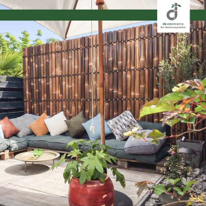 Gemütliche Terrasse mit Sitzlounge aus Paletten und vielen Kissen unter einem Sonnenschirm, umgeben von einer dunklen Bambus-Sichtschutzwand; davor stehen mehrere Pflanzen in Töpfen sowie ein niedriger Beistelltisch auf Holzdeck; oben rechts das DE-COmmerce Logo.