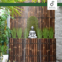 Zen-inspirierter Garten mit einer kleinen Buddha-Statue vor einer dunklen Bambuswand, über die Wasser sanft nach unten fließt. Umgeben von grünen Pflanzen, Palmenblättern und Gräsern entsteht eine ruhige, entspannende Atmosphäre mit weißen Kieselsteinen im Vordergrund; oben rechts das DE-COmmerce Logo.