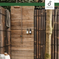 Außendusche im Garten mit Holzlattenwand, freiliegender Metall-Duscharmatur und rundem Duschkopf. Die Seiten sind mit dunklen Bambuselementen aus Bambuslatten und Palmseil verkleidet, links hängt ein Handtuch, rechts steht ein Baumstamm, und an der Wand sind zwei Seifenspender befestigt; oben rechts das DE-COmmerce Logo.