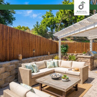 Gemütliche Outdoor-Lounge auf einer Terrasse mit hellem Rattan-Sofa, dekorativen Kissen und Couchtisch, umgeben von einem hohen Bambuszaun, unter einer weißen Pergola mit integriertem Grillbereich und üppiger Begrünung im Hintergrund; oben rechts das DE-COmmerce Logo.